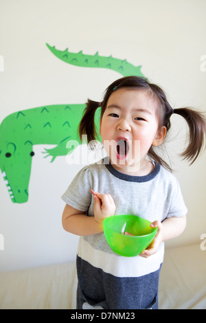 Coreano toddler americano in pig-tail grida alla telecamera di mangiare spuntini dal recipiente verde e coccodrillo wall art dietro. Foto Stock