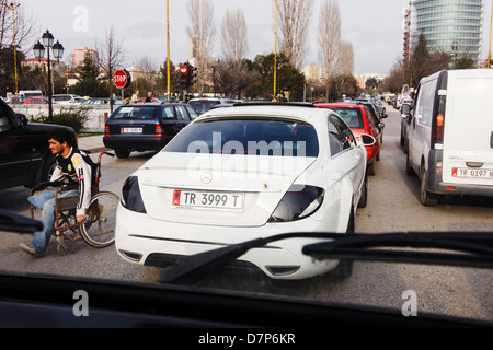 Uomo disabili su sedia a rotelle passando attraverso un inceppamento di traffico con vetture di lusso nel centro di Tirana, Albania Foto Stock