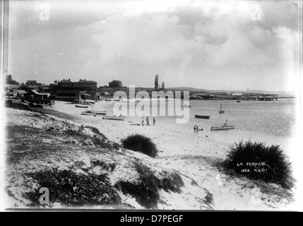 Una fotografia in bianco e nero dell'area di la Perouse nella Frenchmans Bay, con una spiaggia, barche, edifici e persone sulla riva. L'immagine evidenzia il tranquillo paesaggio, con nuotatori e piante visibili lungo la costa, catturando un momento nella storia della zona. Foto Stock
