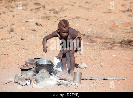 Giovane ragazzo Himba la cottura di cibo sul fuoco aperto nel suo villaggio, Kunene Regione (formerly Kaokoland) nel nord della Namibia Foto Stock