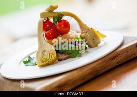 Bistecca di controfiletto pasto servito con carciofi, anelli di cipolla fresca e pomodori a grappolo in una piastra bianca su un blocco di legno Foto Stock