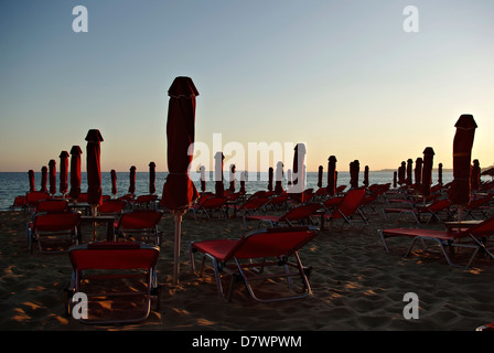 Rosso di ombrelloni e lettini in spiaggia sabbiosa a estate tramonto con il blu del mare e del cielo in background. Foto Stock