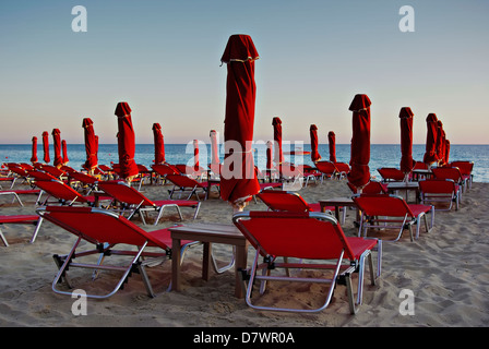 Rosso di ombrelloni e lettini in spiaggia sabbiosa a estate tramonto con il blu del mare e del cielo in background. Foto Stock