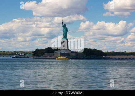 La Statua della Libertà su Liberty Island, New York, NY, STATI UNITI D'AMERICA, con un luminoso giallo taxi acqueo passando davanti ad esso. Foto Stock