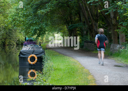 Silenzioso, Scenic, rurale, viale alberato di tratto di strada alzaia con barche ormeggiate & man jogging passato - Leeds Liverpool Canal, Saltaire, nello Yorkshire, Inghilterra, Regno Unito. Foto Stock