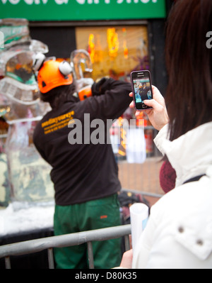 Donna tenendo iphone foto di due uomini carving un blocco di ghiaccio in un pupazzo di neve Scultura su Rhyl town center high street Foto Stock