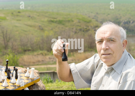 Anziani uomo seduto a giocare a scacchi all'aperto tenendo in mano un pezzo degli scacchi in mano mentre guardando la telecamera Foto Stock