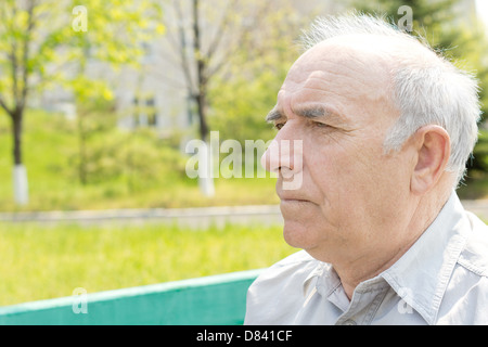 Considerato grave senior uomo seduto lateralmente alla telecamera fissando in lontananza, la testa e le spalle ritratto in un parco rurale. Foto Stock