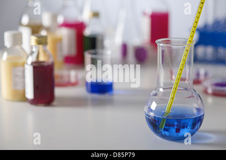 La scienza delle attrezzature di laboratorio con bulbo di vetro in provetta in primo piano contenente sostanze chimiche blu e termometro su un banco di lavoro. Foto Stock