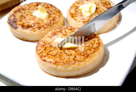 Diffusione di burro con un coltello su un caldo crumpet tostato per colazione o tè Foto Stock