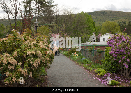 Una donna che spinge una sedia a rotelle attraverso Shelburne Falls ponte di fiori in primavera con la città di Buckland in background. Foto Stock