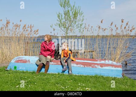 Una madre e il suo giovane figlio e il loro cane seduto su una barca accanto al lago, Schaalsee Zarrentin, Meclemburgo-Pomerania Occidentale, Germania Foto Stock