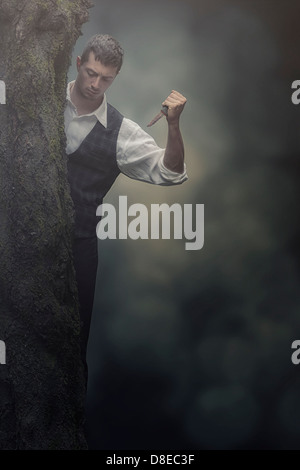 Un uomo nascosto dietro un albero con la lama di un coltello Foto Stock