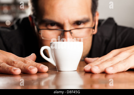 Un uomo che indossa occhiali con bordo nero si appoggia in avanti su un tavolo di legno, il suo viso vicino a una tazza di caffè in ceramica bianca. Le sue mani poggiano in piano sul tavolo su entrambi i lati della tazza, le dita si spalmano. Lo è Foto Stock