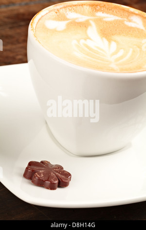 Una tazza in ceramica bianca riempita di latte con un'intricata latte art a forma di foglia sulla superficie, posizionata su un piattino bianco coordinato. Accanto alla tazza, si appoggia una piccola caramella di cioccolato a forma di fiore Foto Stock