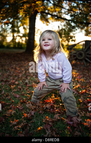 Gentile bambino in piedi su una coperta di foglie di autunno con il sole che tramonta dietro un grande albero. Foto Stock