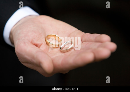 Oro due anelli di nozze tenuto nel palmo aperto di una mano di un uomo che mostra bracciale bianco contro abito scuro Foto Stock