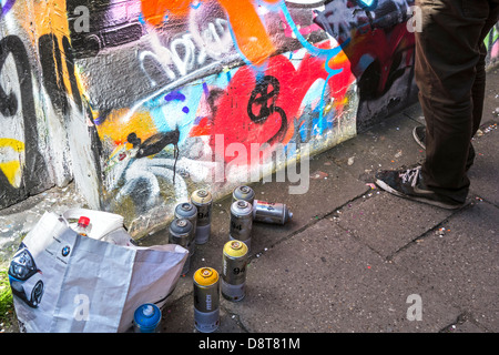 Le bombolette spray sul terreno e giovane la spruzzatura coloratissimo graffito sulla parete di edificio in città Foto Stock