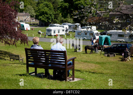 Viaggiatori zingari; coppia seduta fuori sul verde villaggio; case rimorchio nel villaggio di Bainbridge, Richmondshire, North Yorkshire, Regno Unito. 4 giugno 2013. I residenti locali osservano l'accampamento della comunità itinerante lungo il tragitto per la fiera ippica Appleby di Cumbria. La Fiera è un raduno annuale di zingari e viaggiatori che si svolge la prima settimana di giugno, e ha avuto luogo dal regno di Giacomo II, che ha concesso una carta reale nel 1685 consentendo una fiera del cavallo 'vicino al fiume Eden'. Foto Stock