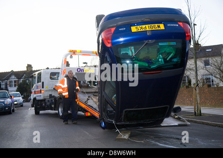 Auto veicolo recupero uomo & carroattrezzi rimuove capovolto auto che ha perso il controllo / si è schiantato in UK tranquilla strada residenziale / road. Foto Stock