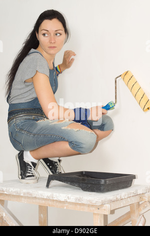 Donna pittura la sua casa con un rullo rannicchiato in giù su un traliccio di legno guardando sopra la sua spalla con un espressione seria guardare qualcosa fuori il telaio a sinistra. Foto Stock