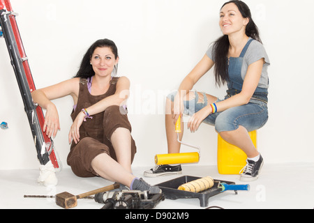 Due attraenti giovani femmine decoratori di sorridere quando si prendono una pausa e rilassarsi sul pavimento ai piedi della scala circondato da apparecchiature di verniciatura. Foto Stock