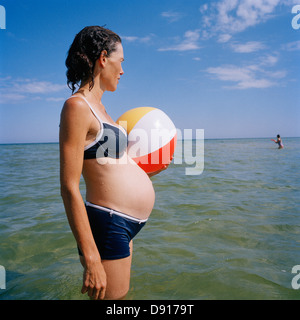 Una donna in stato di gravidanza tenendo una palla spiaggia in piedi in acqua, Svezia. Foto Stock