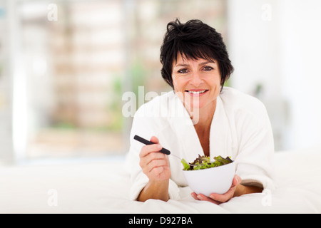 Bella donna matura giacente sul letto e mangiare insalata verde Foto Stock