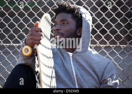 Un giovane serio appoggiata contro una catena collegamento recinto tenendo uno skateboard Foto Stock