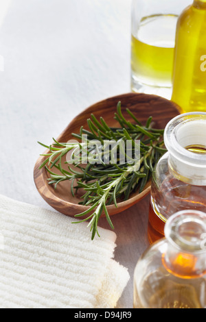 Rametto di rosmarino in una ciotola, erbe, un asciugamano e bottiglie di olio essenziale per aromaterapia Foto Stock