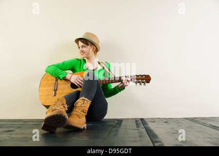 Ritratto di ragazza adolescente seduto sul pavimento, indossando un cappello e suonare la chitarra acustica, Studio shot su sfondo bianco Foto Stock