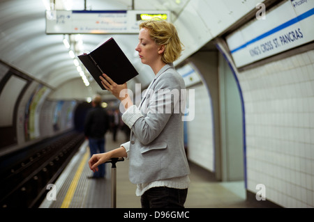 Una donna che legge un libro mentre è in attesa per un metro di Londra treno tubo sulla piattaforma di Kings Cross St Pancras. Foto Stock