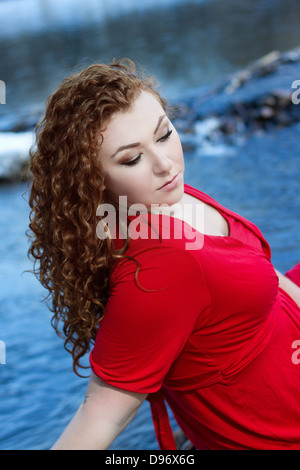 Bella giovane donna con ricci capelli rossi e abito rosso sguardi verso il basso con Vivid Blue River in background Foto Stock