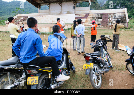 Crossbow Contest Vieng Xai Laos // VIENG XAI, Laos - gli spettatori studenteschi si allineano sui loro scooter e motociclette per assistere a una gara di balestra che si svolge su un campo di gioco sterrato nel terreno accidentato vicino a Vieng Xai nel nord-est del Laos. Il tradizionale evento sportivo attira partecipanti e spettatori dai villaggi circostanti in questa regione rurale montana. Vieng Xai, situata nella provincia di Houaphanh, è conosciuta per il suo paesaggio carsico calcareo e la rete di grotte che servirono come quartier generale per il Pathet Lao durante la seconda guerra d'Indocina. La città si trova vicino al confine con il Vietnam in una delle zone di L Foto Stock