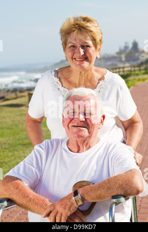 Disabili uomo senior essendo spinto sulla sedia a rotelle dalla sua amorevole moglie alla spiaggia sulla giornata di sole Foto Stock