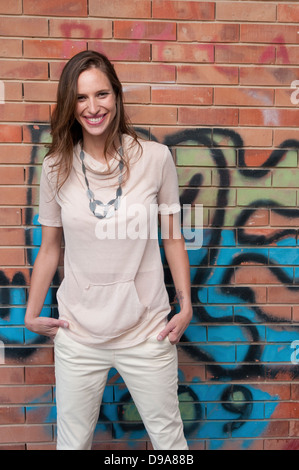 Un sorridente elegante e alla moda giovane donna sta in piedi di fronte ad un muro di mattoni con graffiti modello di rilascio disponibili Foto Stock