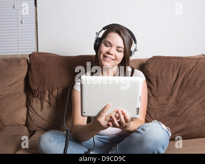 Bella bruna giovane donna che indossa jeans e una maglietta di casa utilizzando il suo tablet pc e le cuffie di grandi dimensioni. Foto Stock