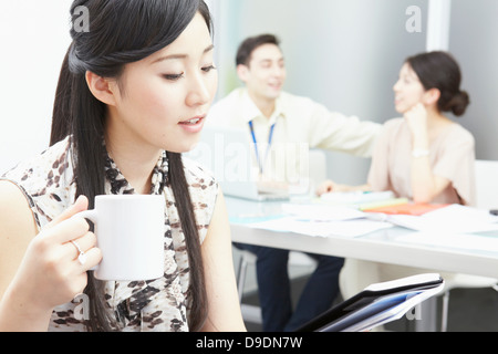 Donna che mantiene la tazza, colleghi in background Foto Stock