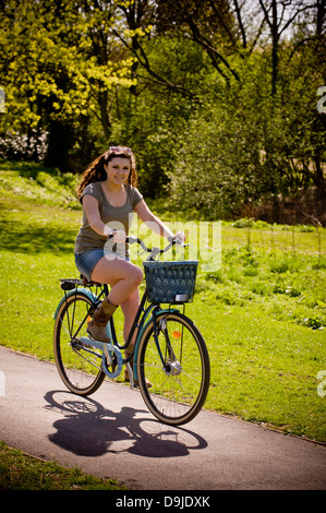 Giovane donna caucasica, vestita casualmente in una breve gonna di jeans che guida una bicicletta tradizionale in stile shopper attraverso un parco del Regno Unito. Foto Stock