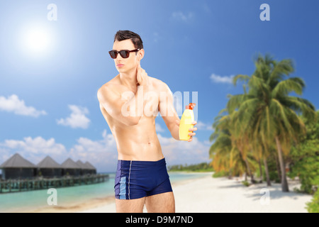 Bel ragazzo mettendo in crema solare, su di una spiaggia di sabbia con palme e acqua villa cottages Foto Stock