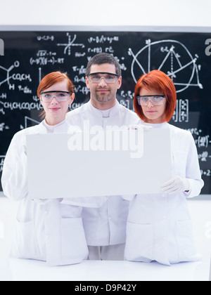 Close-up di due belle ragazze studenti e un insegnante in un laboratorio di chimica sorridente e tenendo in mano un banner di vuoto e un bla Foto Stock