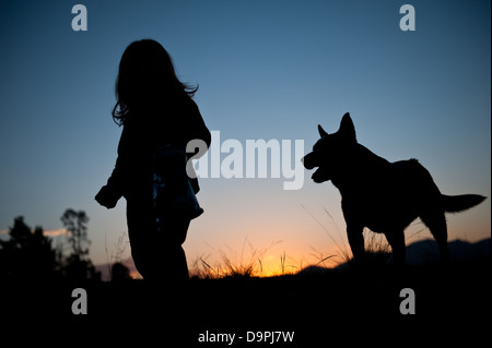 Bel tramonto della silhouette toddler giovane ragazza con i capelli ondulati camminando nel campo con il suo cane strettamente dietro di lei. Foto Stock