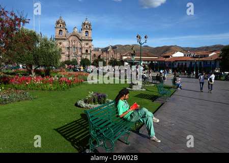 Backpacker seduto sulla panchina a leggere un libro, la chiesa di Compañia de Jesus sullo sfondo, Plaza de Armas, Cusco, Perù Foto Stock
