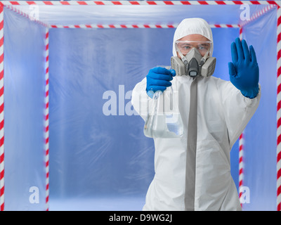 Persona in biohazard tuta protettiva che indossa una maschera di respirazione chiamando un arresto sollevando una mano mentre si tiene una bottiglia trasparente di liquido incolore in altri in piedi di fronte ad una tenda di contenimento Foto Stock