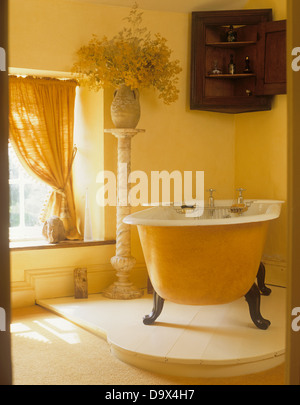 Artiglio bagno piede sul pavimento sopraelevato in bagno di colore giallo con antichi angoliera con vaso di fiori su alto piedistallo Foto Stock