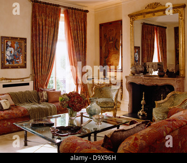 Rosso divani modellato rivolta tabella di caffè di vetro davanti al camino sotto antico specchio dorato in sala da disegno con drappi rossi Foto Stock