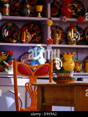 Rosso + arancio sedia al semplice tavolo in legno nella parte anteriore della collezione di ceramiche colorate su ripiani viola Foto Stock