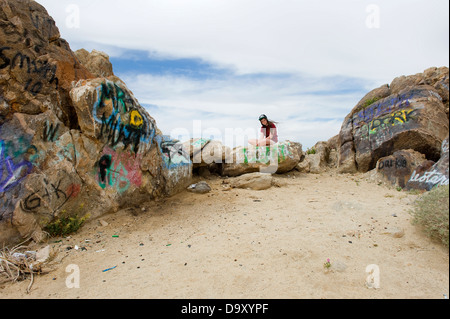 Ragazza adolescente in 'OBAY' cap seduti sulla coperta di graffiti rocce nel deserto di Mojave, California, Stati Uniti d'America. Foto Stock