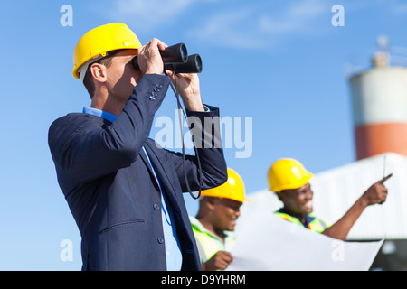 Architetto usando il binocolo guardando il sito in costruzione con lavoratori edili Foto Stock