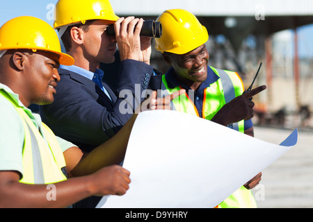 Project manager utilizzando il binocolo cercando sul sito di costruzione con i lavoratori Foto Stock